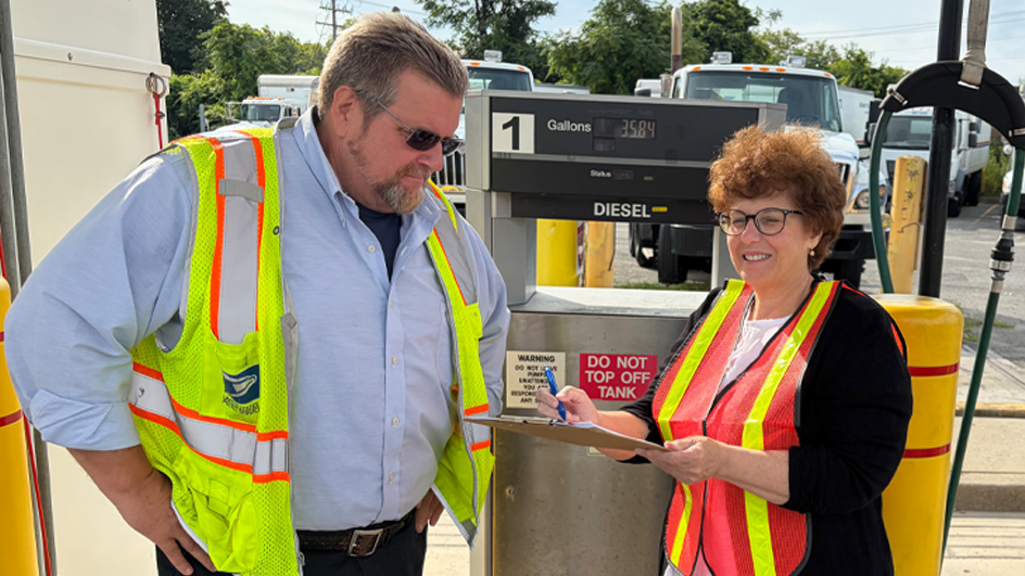 A man and a woman wearing bright-colored safety vests standing side-by-side in front of a fuel pump