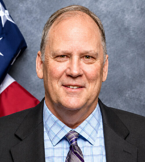 Professional-looking headshot of a man wearing a black suit jacket, light blue shirt and dark-colored tie with an American flag in the background