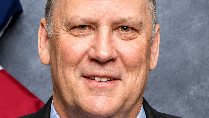 Professional-looking headshot of a man wearing a black suit jacket, light blue shirt and dark-colored tie with an American flag in the background