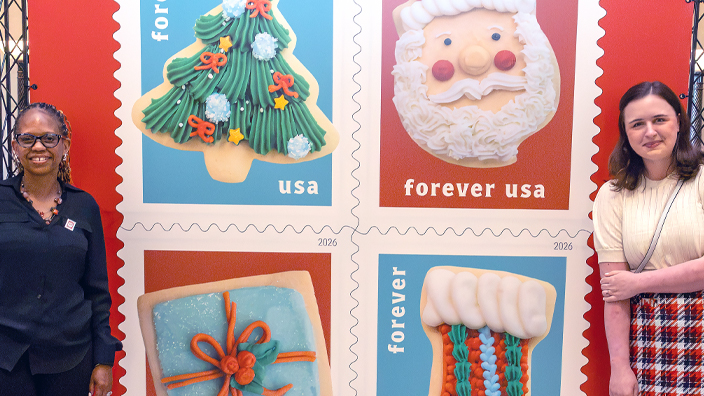 Two women standing on either side of an enlarged image of the USPS Christmas Cookies stamps featuring photos of cookies decorated like a Christmas tree, the face of Santa Claus, a stocking and a wrapped gift.