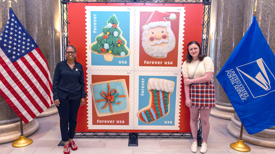 Two women standing on either side of an enlarged image of the USPS Christmas Cookies stamps featuring photos of cookies decorated like a Christmas tree, the face of Santa Claus, a stocking and a wrapped gift.
