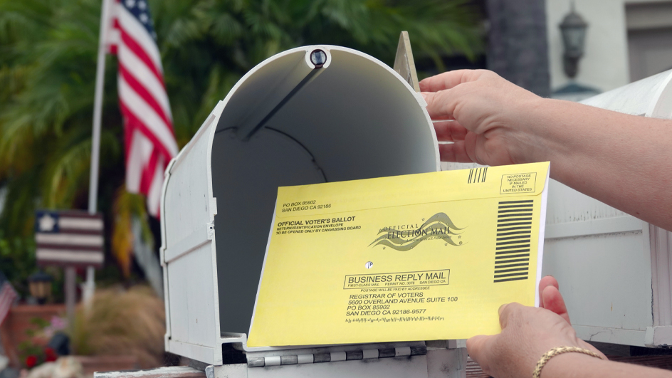 Closeup of a yellow envelope with the Election Mail logo being put into a mailbox