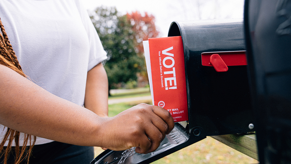 A woman removing a piece of mail that reads “VOTE!” from a black, curbside mailbox