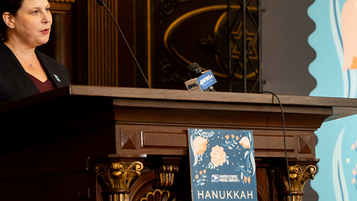 A woman in a dark suit standing behind a wooden podium beside an enlarged image of the Hanukkah stamp
