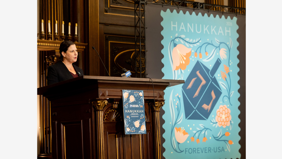 A woman in a dark suit standing behind a wooden podium beside an enlarged image of the Hanukkah stamp