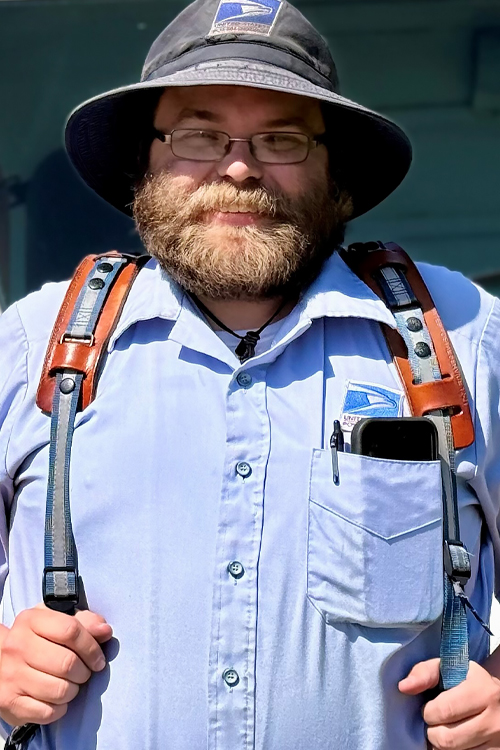 A uniformed USPS employee standing in front of an LLV delivery vehicle and wearing a blue hat and button-up shirt