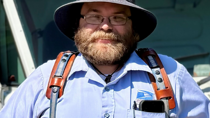 A uniformed USPS employee standing in front of an LLV delivery vehicle and wearing a blue hat and button-up shirt