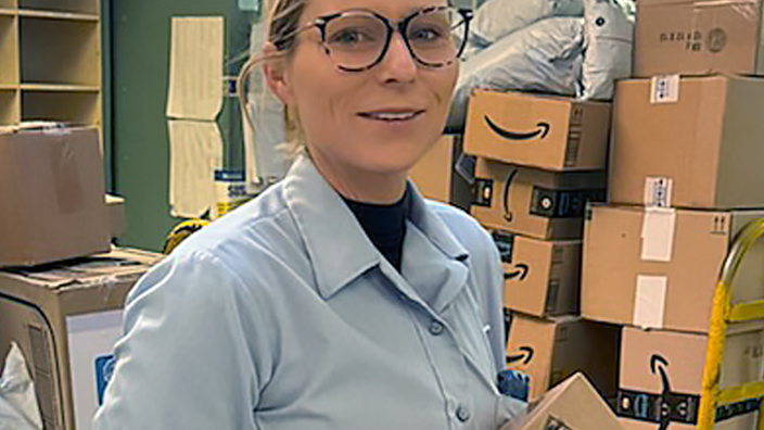 A uniformed USPS employee standing in a Post Office work area and holding a package