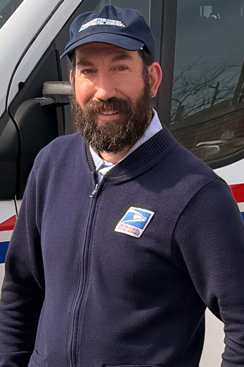 Uniformed USPS employee wearing a dark blue sweater and dark blue hat standing outdoors next to a delivery vehicle