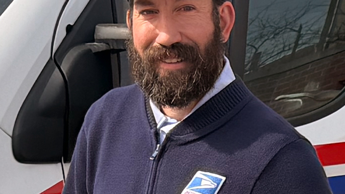 Uniformed USPS employee wearing a dark blue sweater and dark blue hat standing outdoors next to a delivery vehicle
