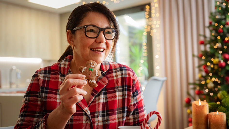 Smiling woman in pajamas eating a gingerbread man cookie and drinking hot chocolate