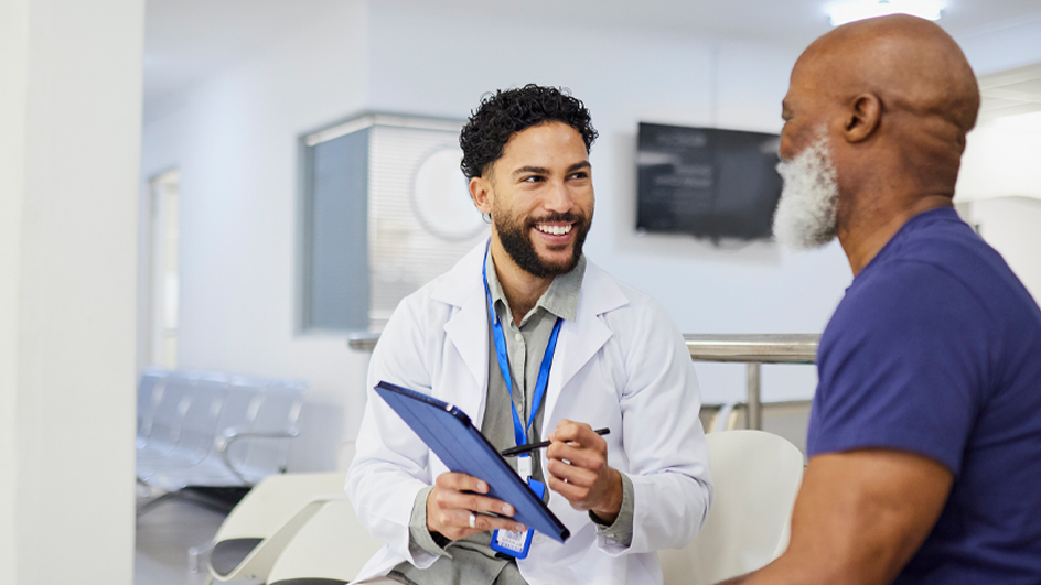 Male doctor wearing eyeglasses and white lab coat and holding a clipboard while consulting with a male patient in a doctor’s office