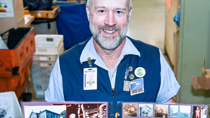 A smiling uniformed USPS employee holds up pages from a scrapbook