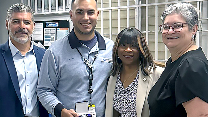 Five smiling USPS employees standing side by side on the workroom floor of a Post Office