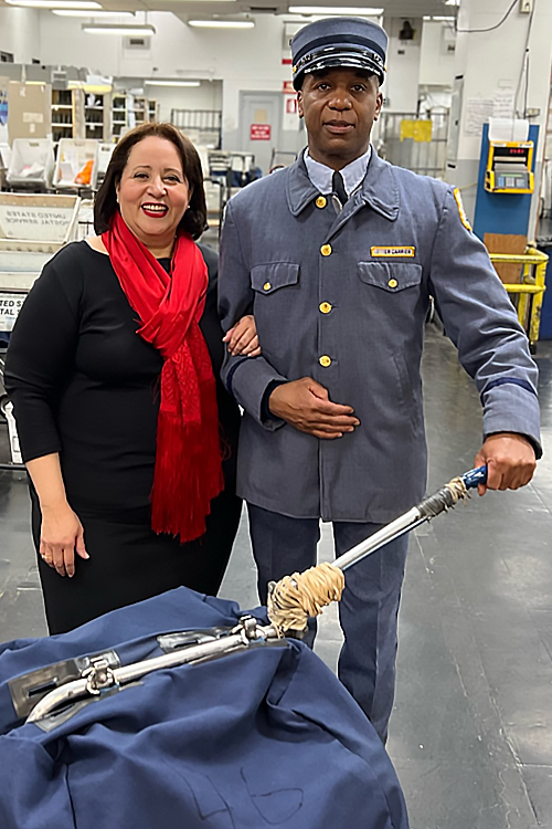 A woman in business attire standing next to a USPS letter carrier wearing a vintage uniform