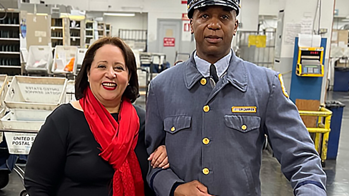 A woman in business attire standing next to a USPS letter carrier wearing a vintage uniform