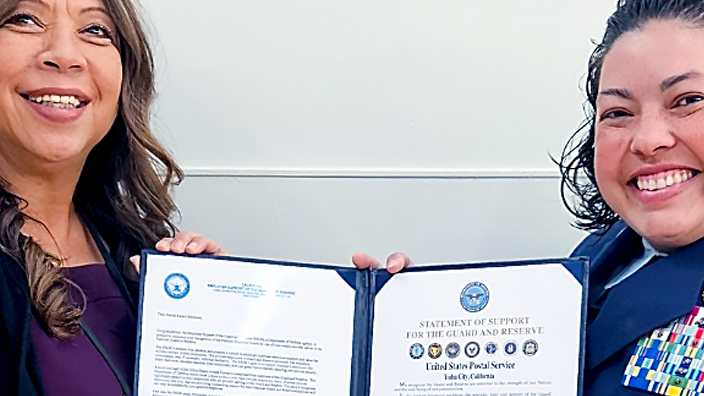 A woman in business attire and a woman in a U.S. Air Force uniform standing side by side and holding up an award