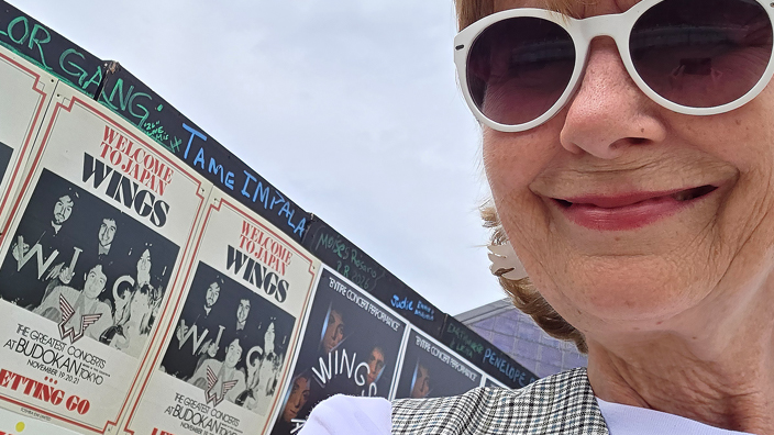 A woman in sunglasses smiles in front of a wall of posters advertising concerts by the pop band Wings.