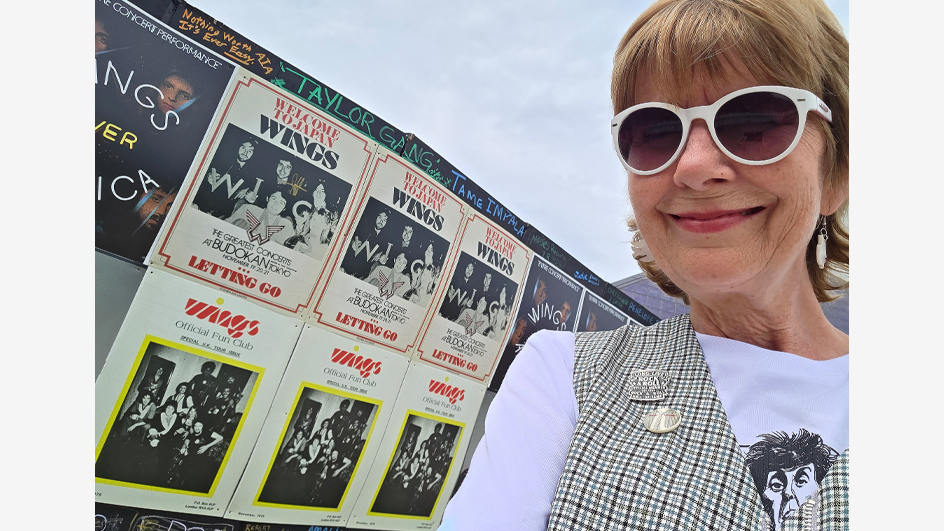 A woman in sunglasses smiles in front of a wall of posters advertising concerts by the pop band Wings.