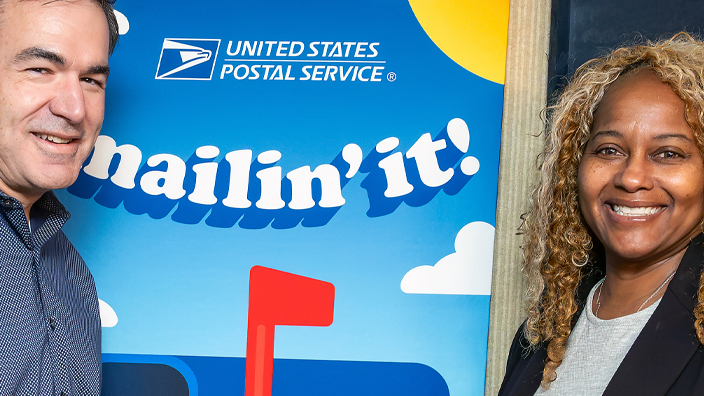 A man and a woman standing on either side of a sign for the “Mailin’ It!” podcast