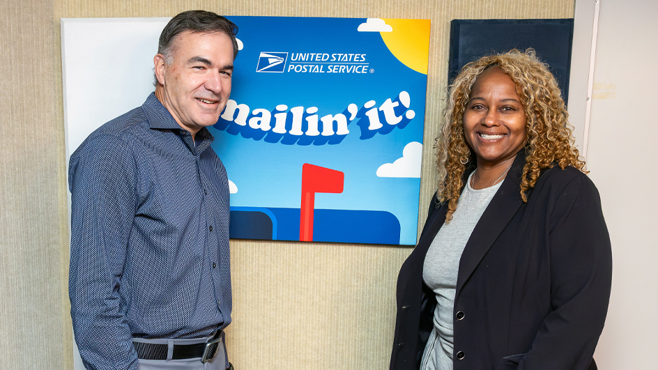 A man and a woman standing on either side of a sign for the “Mailin’ It!” podcast